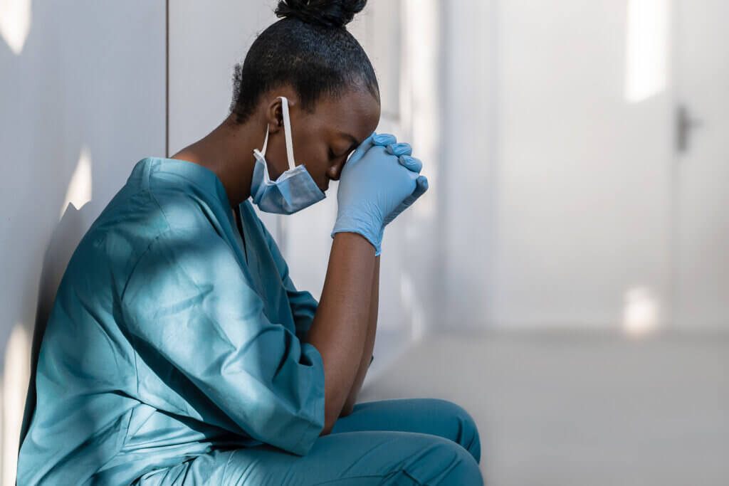 Tired female african nurse wearing face mask sits on hospital floor