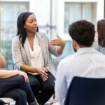 A young female counselor gestures as she sits in a circle with clients